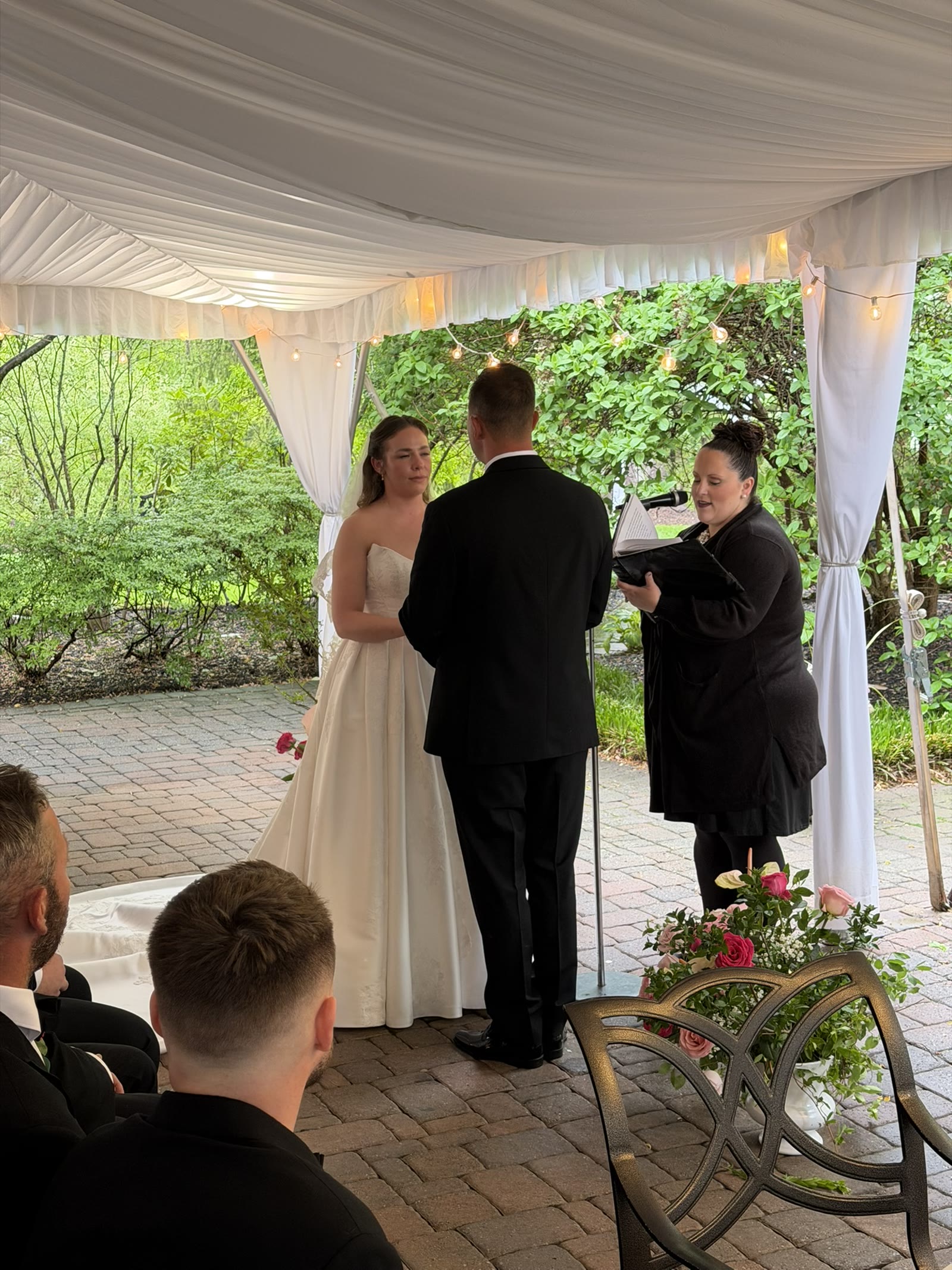 Guests watch an outdoor wedding ceremony in a garden setting.